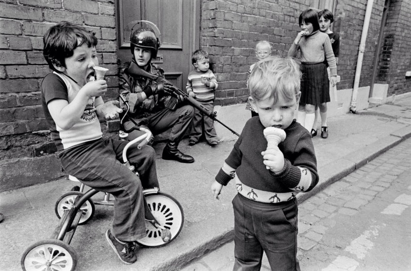 Children eat their ice creams while soldiers patrol the streets of Londonderry 1979