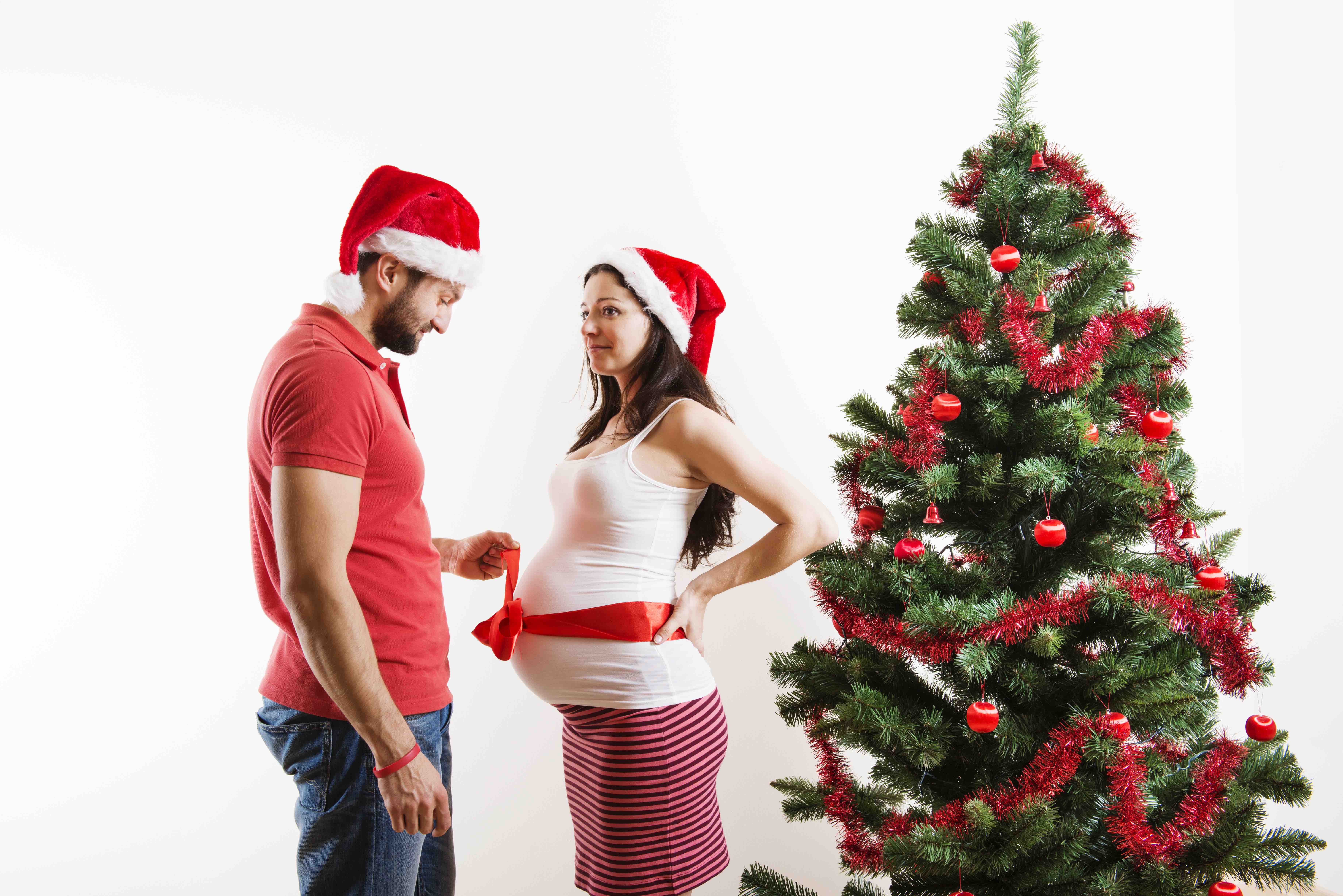 Young couple is standing close to christmas tree. Woman is pregnant and tied with red ribbon.